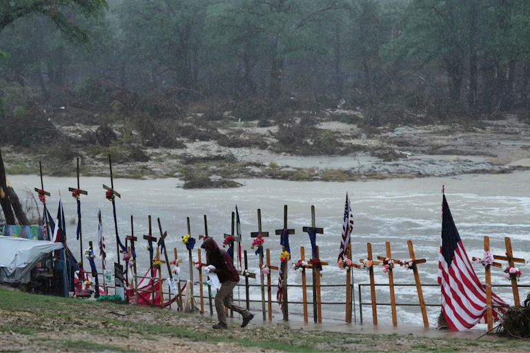 View of the Guadalupe River with crosses and a flag on the banks to honor flood victims and a person in a raincoat walking away in the foreground.