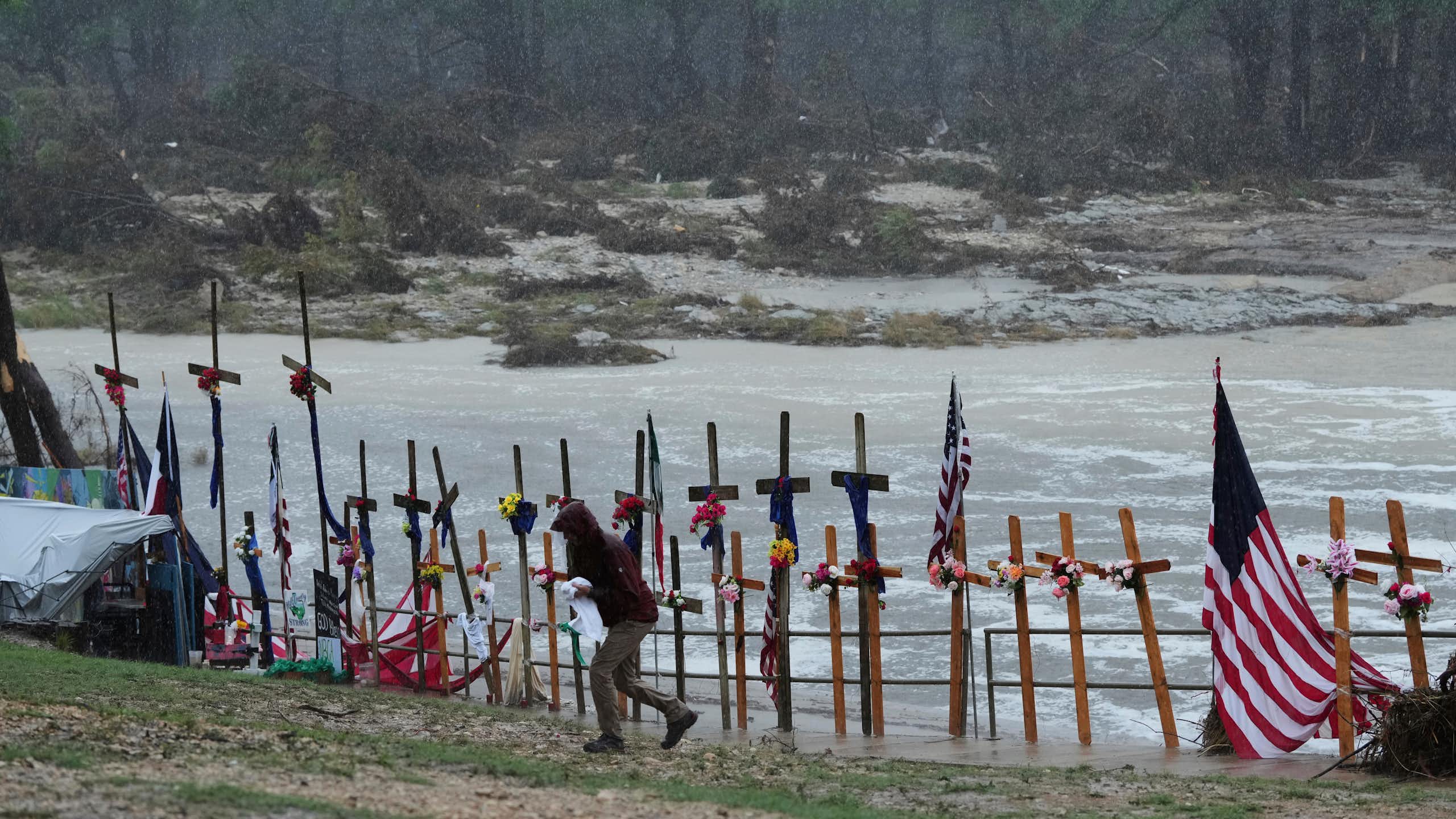 View of the Guadalupe River with crosses and a flag on the banks to honor flood victims and a person in a raincoat walking away in the foreground.
