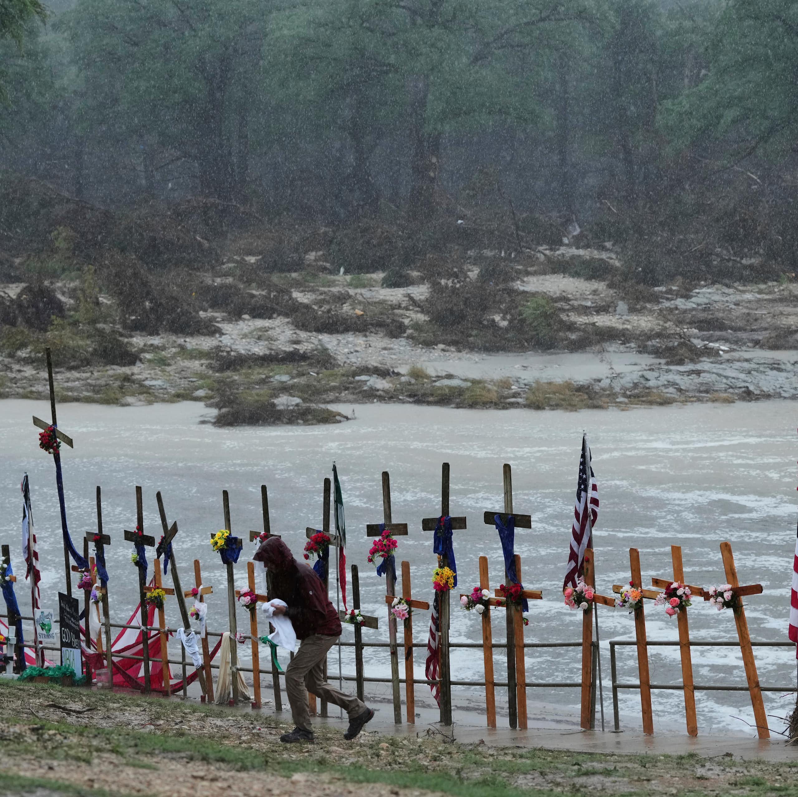 View of the Guadalupe River with crosses and a flag on the banks to honor flood victims and a person in a raincoat walking away in the foreground.