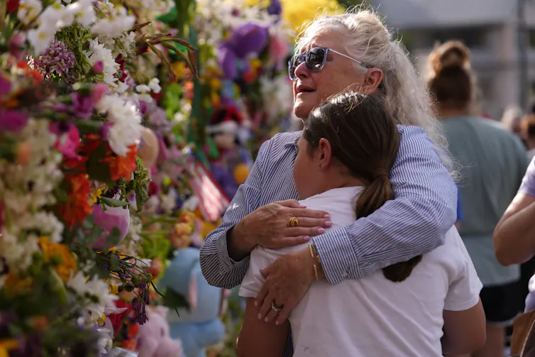 A grandmother embraces a young woman in front of a wall of flowers.