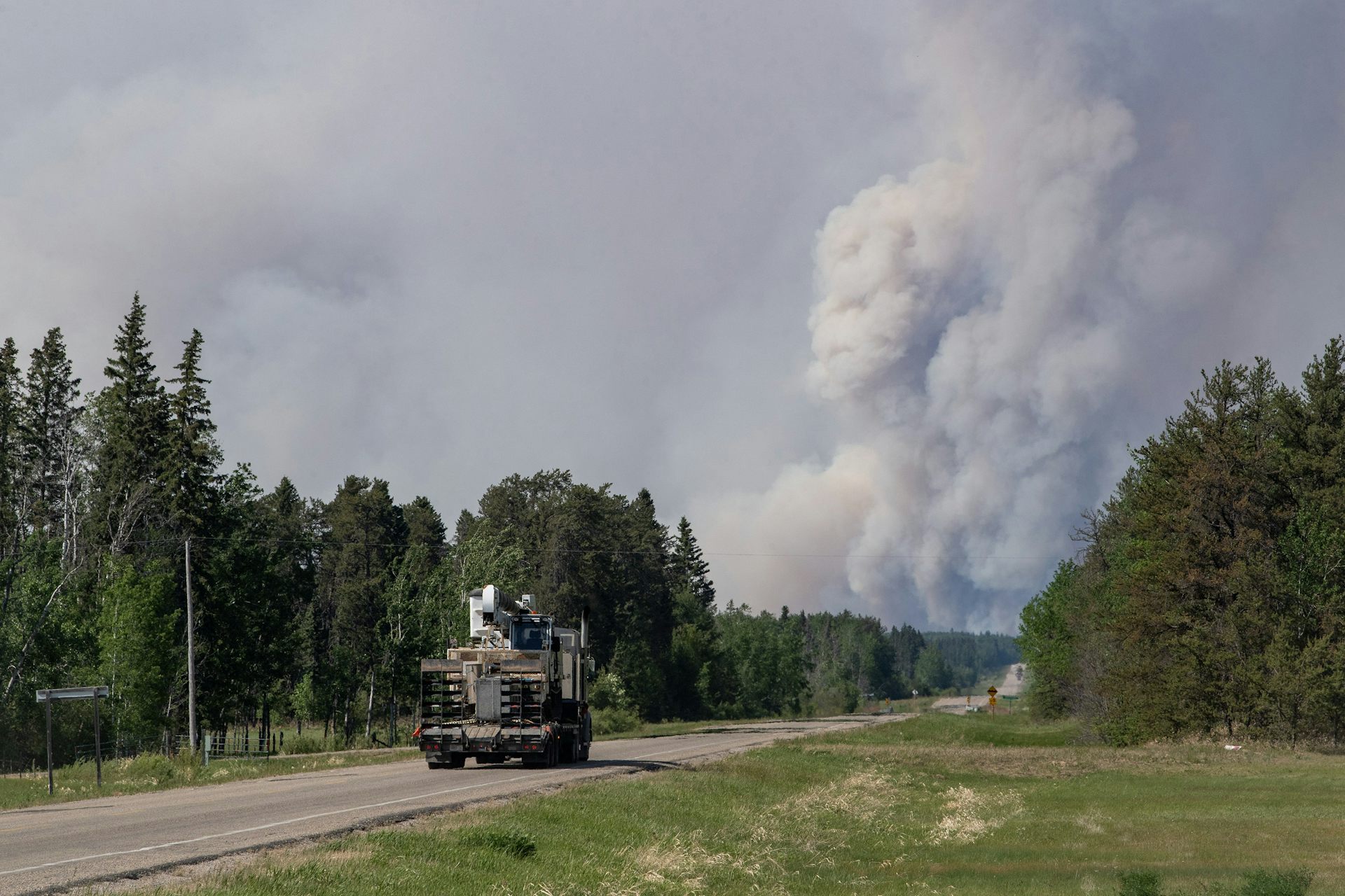 A truck on a highway in a forested area, a large plume of white smoke rises above trees in the distance