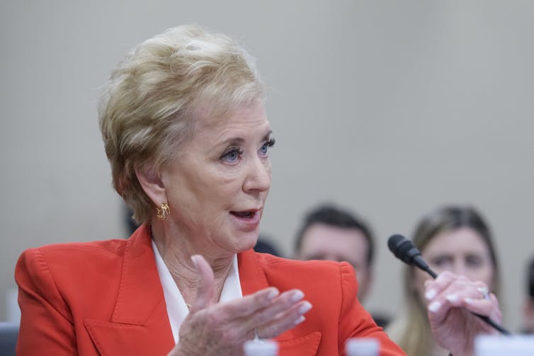 a woman wearing an orange jacket gestures in front of a microphone