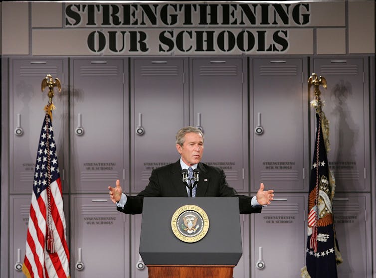George W. Bush speaks against a school locker backdrop, next to an American flag and another flag. Above him are the words 'Strengthening our schools.'
