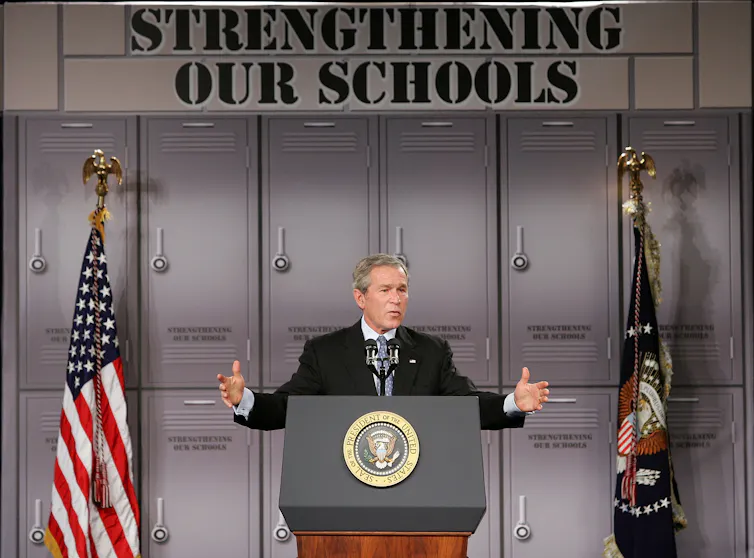 George W. Bush speaks against a school locker backdrop, next to an American flag and another flag. Above him are the words 'Strengthening our schools.'