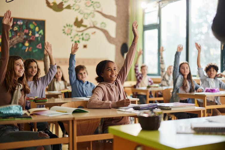 young students sitting at their desks in a classroom raise their hands