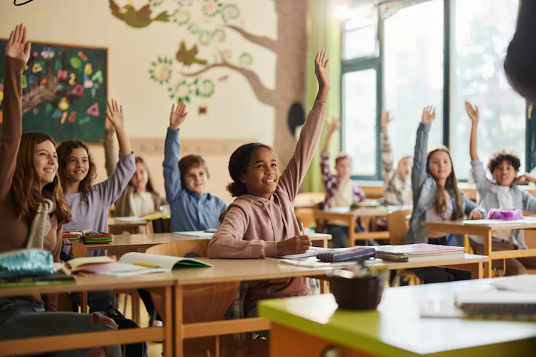 young students sitting at their desks in a classroom raise their hands