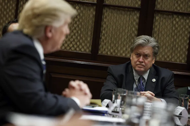 A dishevelled-looking man with shaggy grey hair listens as a rotund man with orange-ish blond hair speaks.