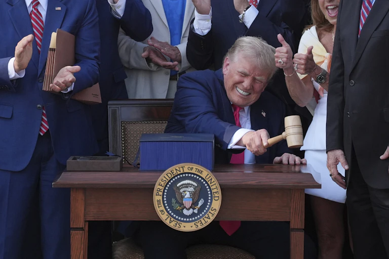 A seated man makes a face and bangs a gavel surrounded by cheering people.