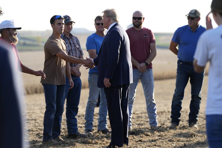 El hombre de Rotunde con un traje azul con cabello blanco naranja se sacude con la mano con un grupo de agricultores en el campo.