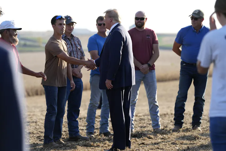 A rotund man in a blue suit with orange-ish white hair shakes hands with a group of farmers in a field.