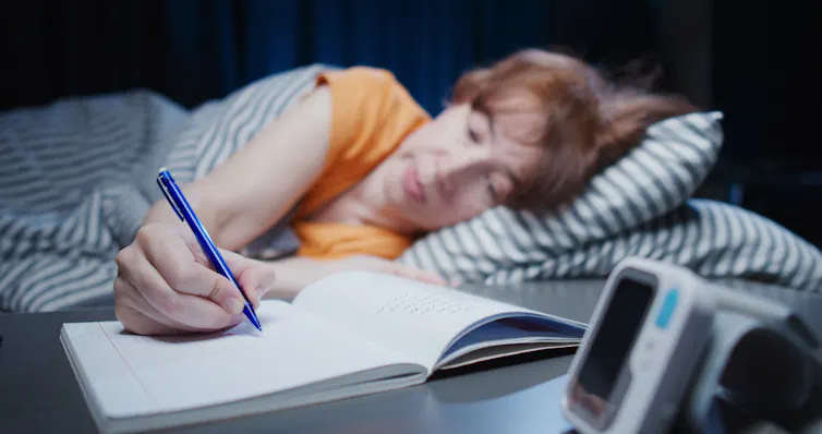 woman lying in bed reaches with pen to write in notebook