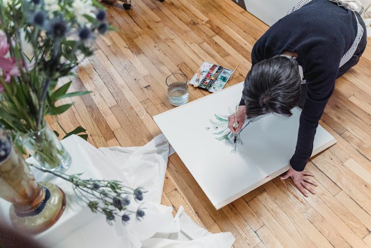 A woman paints flowers on paper while sitting on floorboards