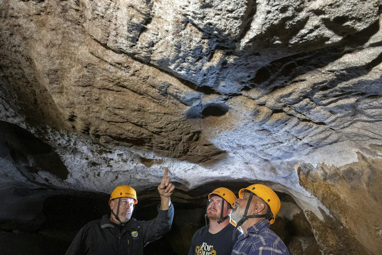 Three men in yellow hard hats admire the ceiling of a cave with deep grooves in it.
