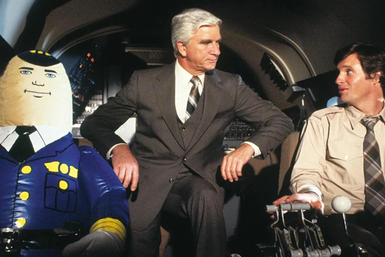 En la cabina de pilotos de un avión, dos hombres en traje conversan al lado de un muñeco hinchable vestido de piloto.