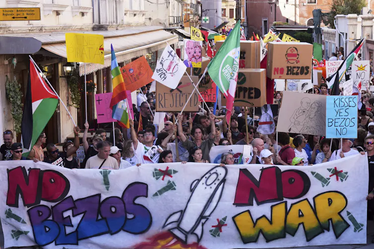 A crowd of protests holding flags, signs and a banner that says 'NO BEZOS NO WAR'