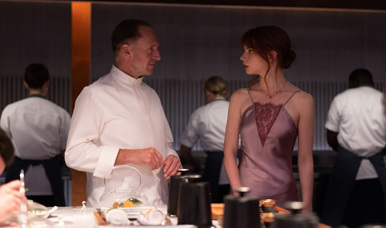 Screencap of a movie showing an older white man in a chef uniform standing beside a young white woman in a slip dress
