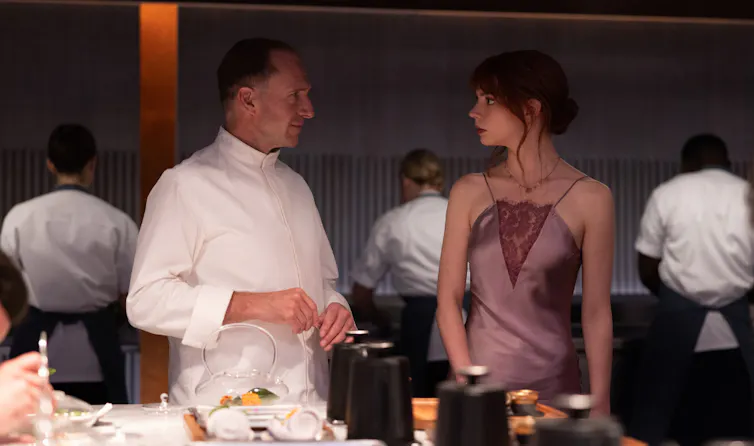 Screencap of a movie showing an older white man in a chef uniform standing beside a young white woman in a slip dress
