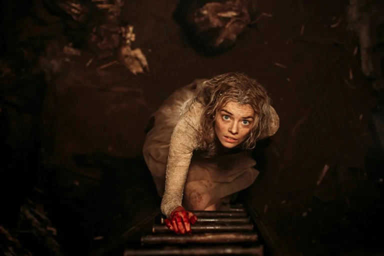 Screenshot of a white woman in a tattered wedding dress looking upwards as she stands at the base of a ladder