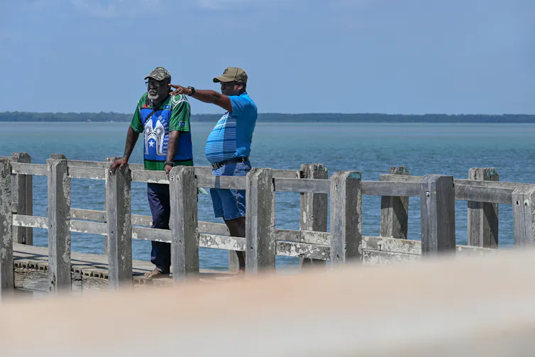 Two men stand on a wharf, one pointing into the distance