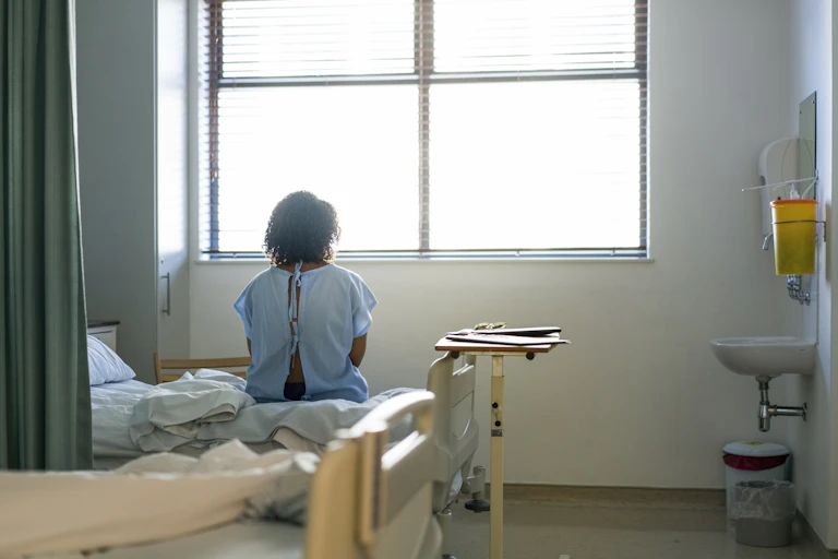 Woman in medical gown sits on bed in hospital facing a window.