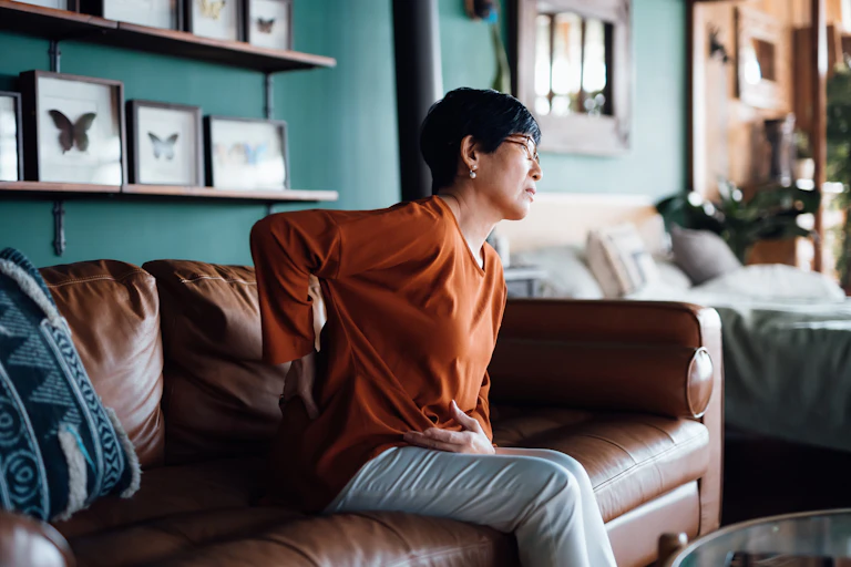 Asian woman sitting on sofa, one hand on lower back, in pain