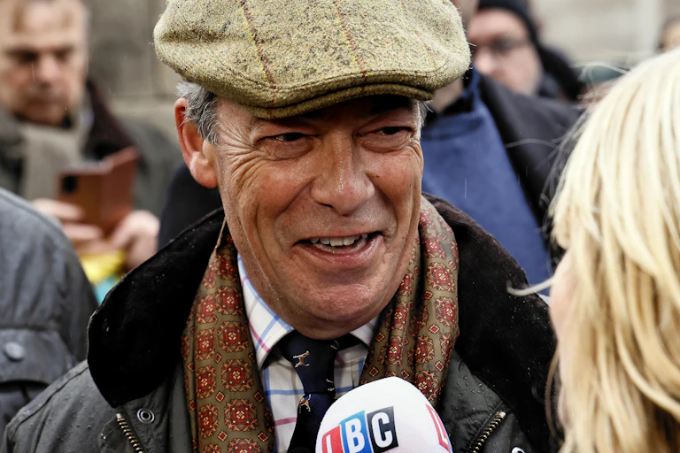 Nigel Farage in a wool cap at a farmer's protest smiles and speaks to an LBC reporter
