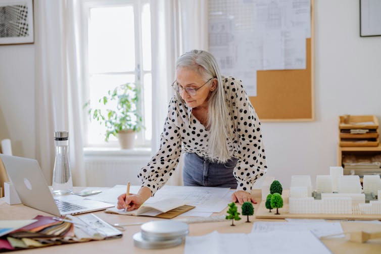 Une femme aux longs cheveux gris penchés sur un bureau, crayon à la main