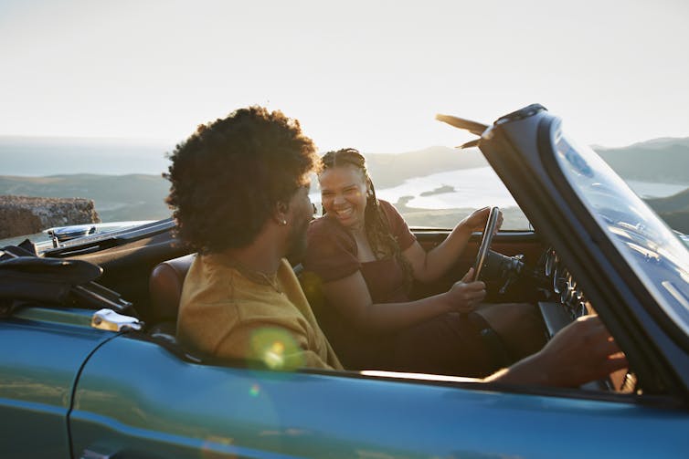 A young woman turns to face a young man while sitting in a convertible car