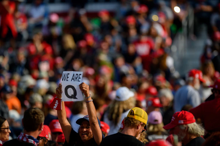 A woman standing in the middle of a large crowd, holding up a 'Q' sign.