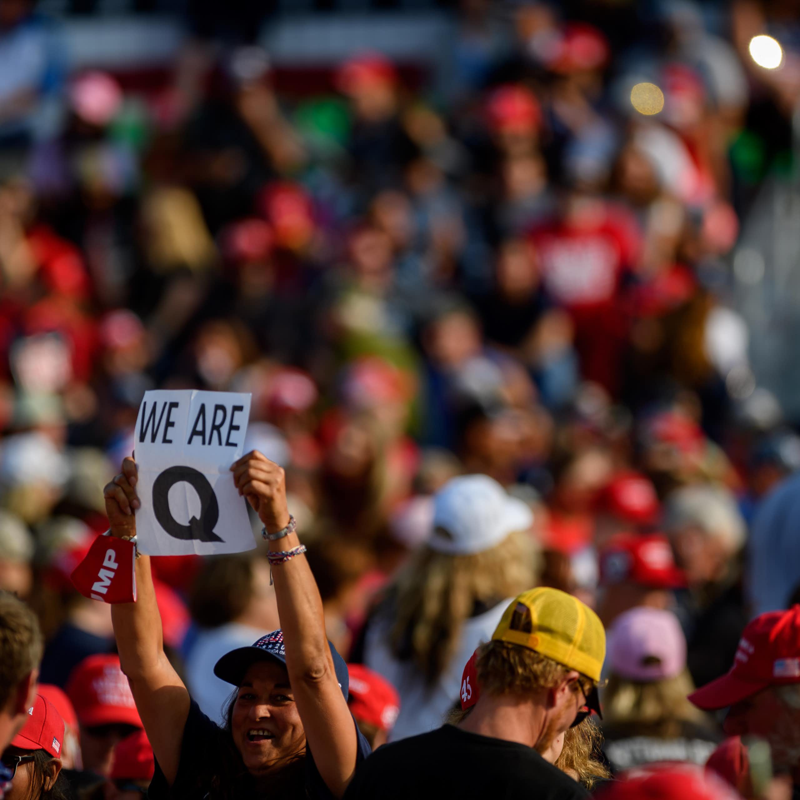 A woman standing in the middle of a large crowd, holding up a 'Q' sign.