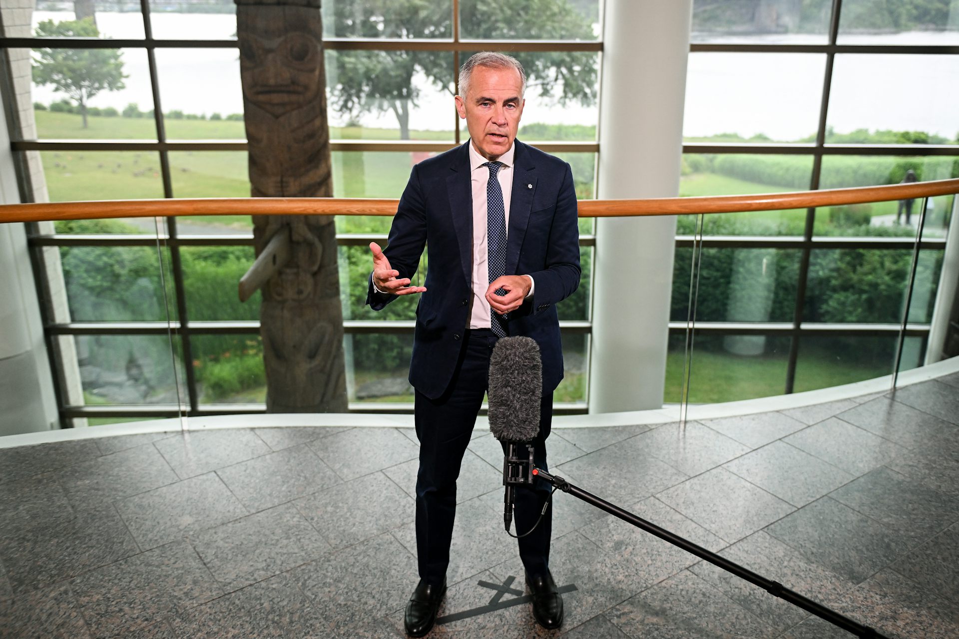 A man with short dark hair in a dark blue suit stands on a stage behind a microphone with windows behind him showcasing trees and greenery.