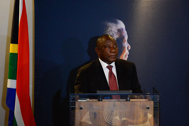 Man in suit and tie at a podium with a flag to one side and the profile of an older man depicted on the wall behind him