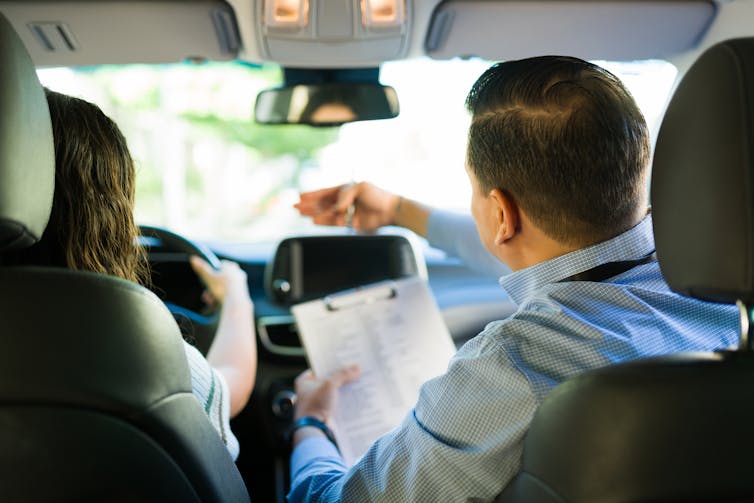 A rear view of an instructor holding a clipboard and guiding a student during a driving lesson.