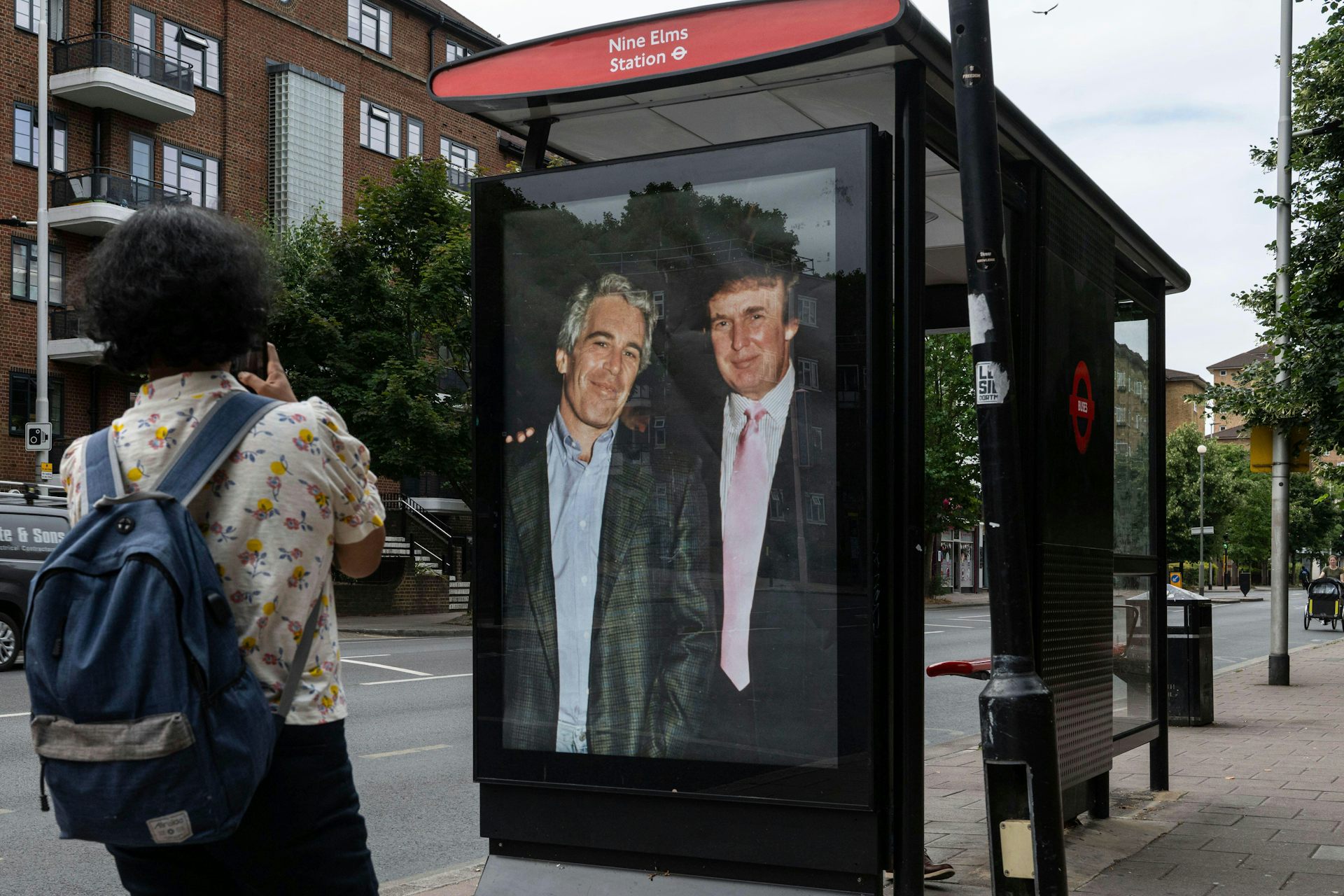 A man with a backpack takes a photo of a bus shelter with a poster showing Donald Trump with Jeffrey Epstein.