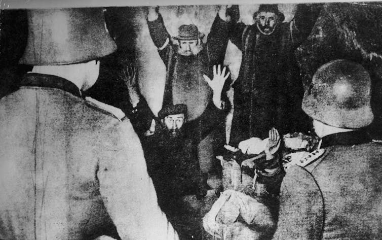 Men are seen looking afraid and with their hands up, looking toward two men with uniforms and helmets, in a faded black-and-white photo.