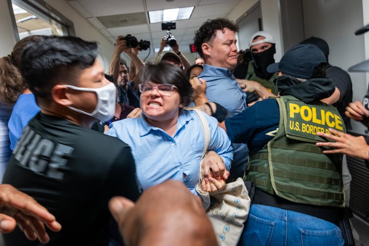 A woman wearing a blue shirt grimaces as she is held back by a man wearing a black shirt that says 'police.' Other people appear to fight alongside them.
