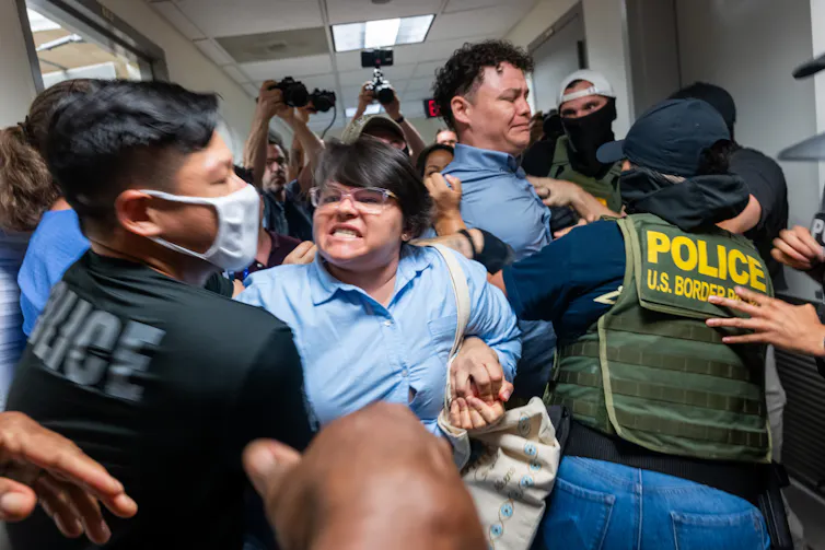 A woman wearing a blue shirt grimaces as she is held back by a man wearing a black shirt that says 'police.' Other people appear to fight alongside them.