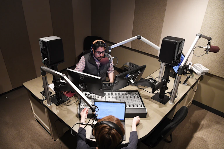 Two people in a radio studio, sitting on opposite sides of a table, with electronic and digital media around them.