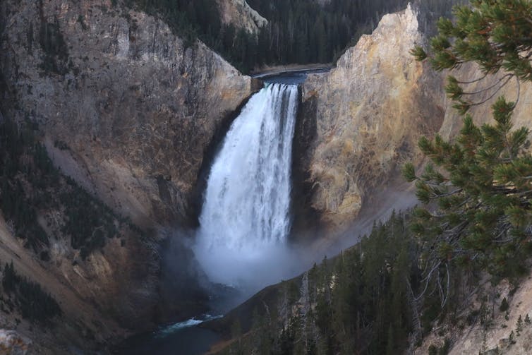 A large waterfall pours over a steep cliff into a river below, surrounded by rocky canyon walls and forested slopes.