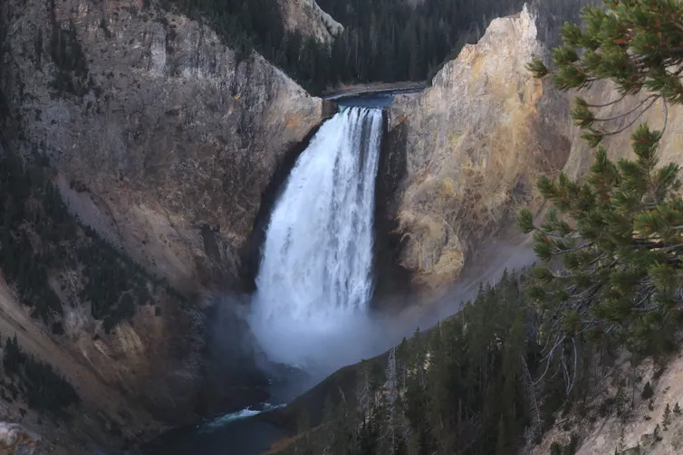 A large waterfall pours over a steep cliff into a river below, surrounded by rocky canyon walls and forested slopes.