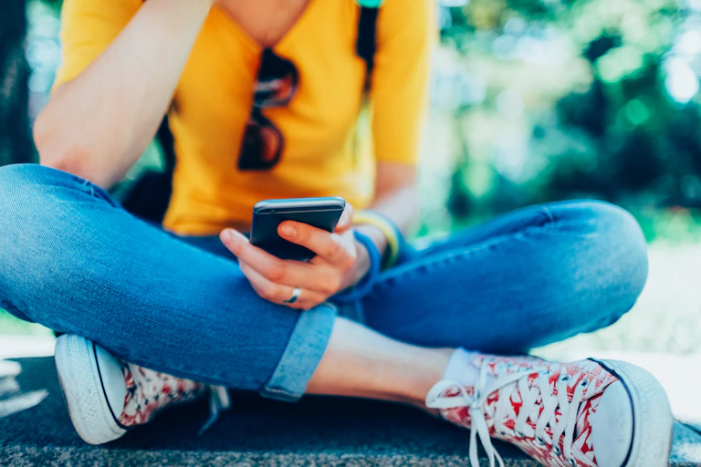 Young woman sitting cross-legged on ground looking down at smartphone in hand