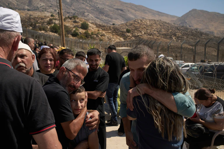 A crowd of people stands near a security fence amid hills, as several people embrace one another.