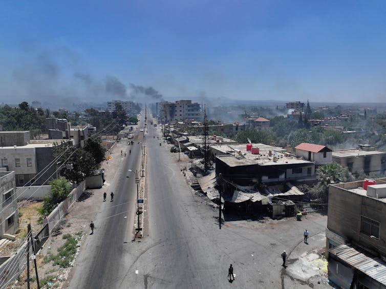 A wide road seen from above, with damaged buildings alongside it and smoke rising against a blue sky.