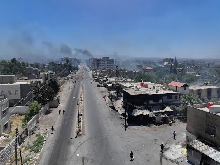 A wide road seen from above, with damaged buildings alongside it and smoke rising against a blue sky.