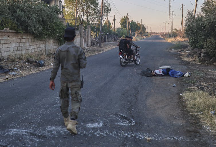 A man in camo walks down a road as two men up ahead on a bicycle look at a covered body on the ground.