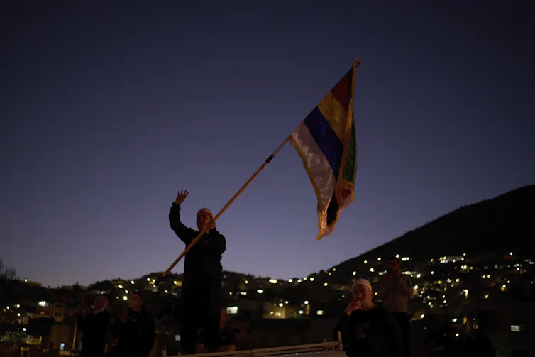 A person holding a brightly colored flag waves as night falls in a hilly area.