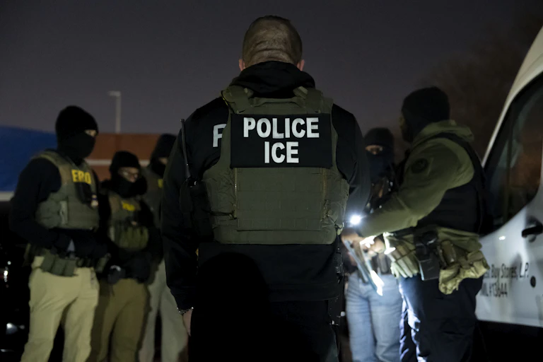 A group of men wearing police vests and dark clothing and face masks stand together outside at nighttime. One wears a vest that says Police ICE.