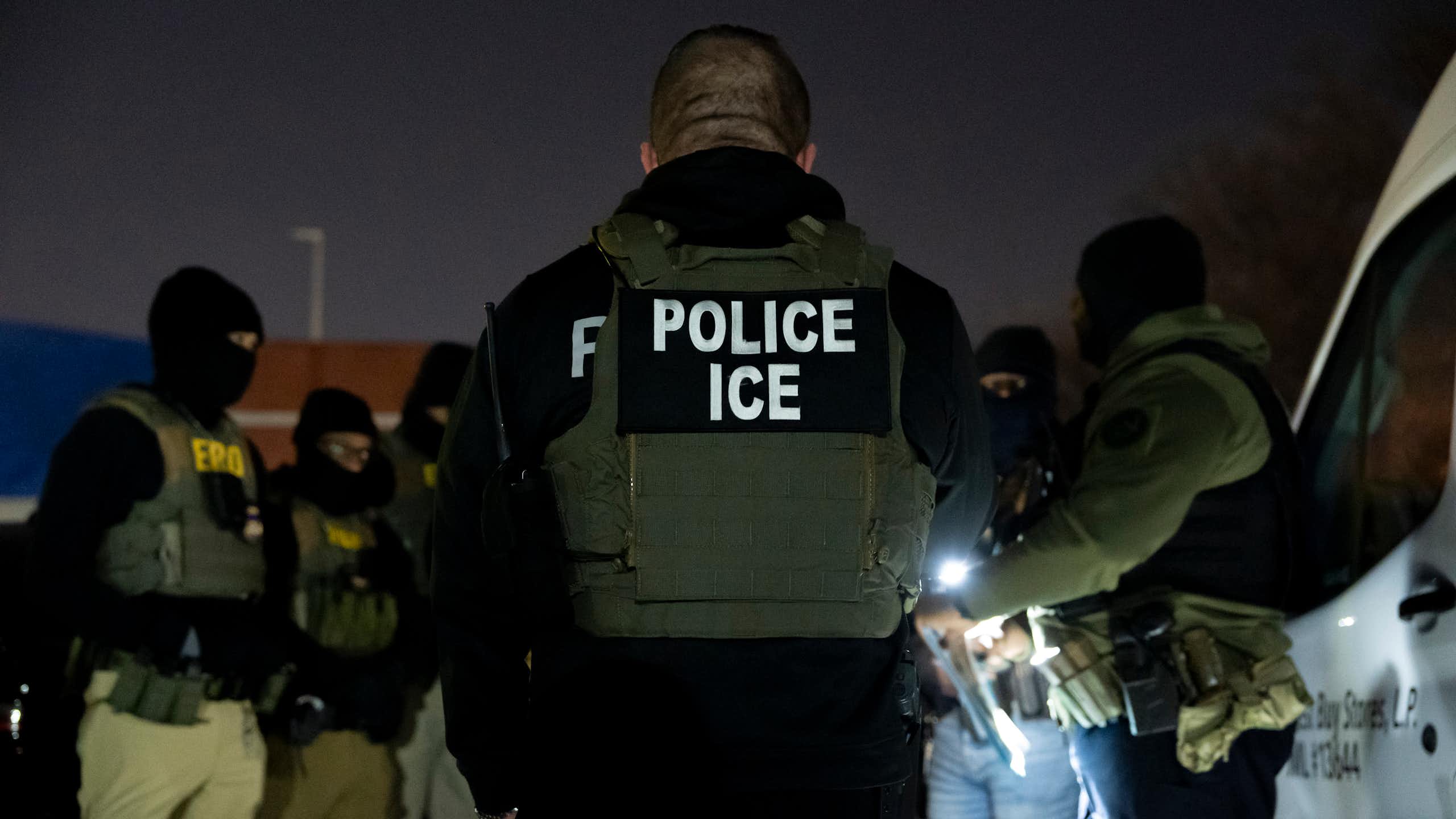 A group of men wearing police vests and dark clothing and face masks stand together outside at nighttime. One wears a vest that says Police ICE.