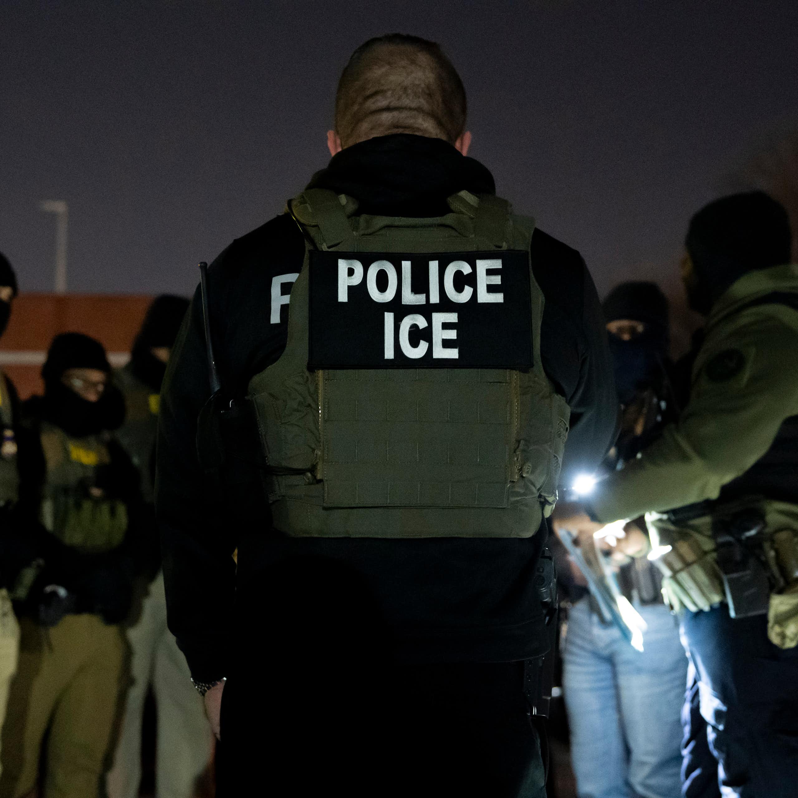 A group of men wearing police vests and dark clothing and face masks stand together outside at nighttime. One wears a vest that says Police ICE.