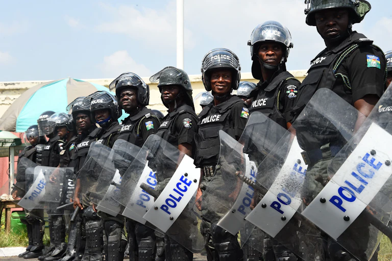 Men in helmets, bulletproof vests and dark uniforms, holding shields labelled Police, standing in line